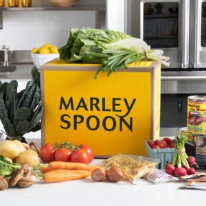 a meal delivery box from Marley Spoon on a white kitchen counter, surrounded by an assortment of vegetables and other grocery items