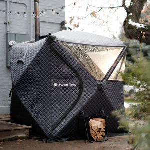 a Sweat Tent setup outside of a grey brick building, with a stack of wood in front and a string of patio lights overhead