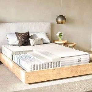 A bed with a cutaway view showing the internal layers of the mattress, including springs and foam. The bed is neatly made with several pillows and a modern, minimalist bedroom setting.