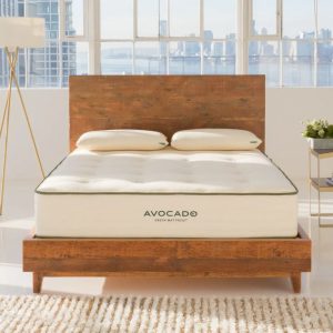 An Avocado Green Mattress in a modern bedroom with a wooden bed frame. The mattress has a white cover with the "AVOCADO" logo. The room features large windows, a standing lamp, and two pillows on the bed.