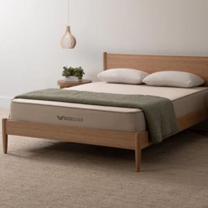 A minimalist bedroom showcasing a Brooklyn Bedding EcoSleep mattress on a wooden bed frame. The mattress is topped with a green blanket and two pillows. A modern pendant light and a small plant on a side table complete the room.