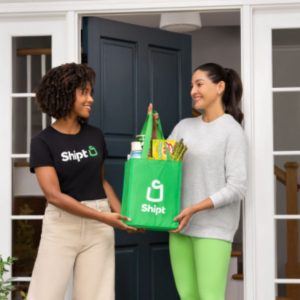 A woman with a "Shipt" shirt hands over a grocery bag to another woman.