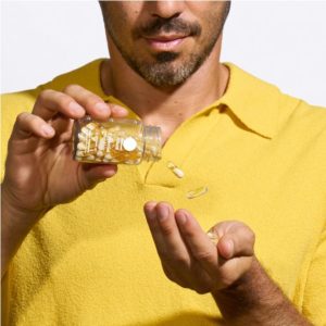 Man in a yellow shirt holding a clear bottle labeled "Ritual Essential for Men," with clear capsules pouring into his hand.