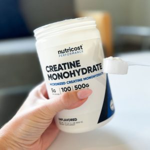 a professional product tester holding a medium sized container of nutricost creatine monohydrate next to a scoop for size comparison