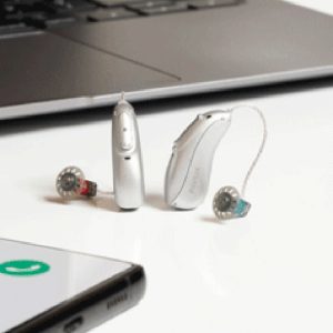 A pair of Phonak hearing aids placed upright on a white surface, with a laptop in the background. The hearing aids are silver with transparent ear tips, and the Phonak logo is visible on the devices.