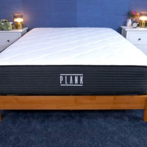 Plank Firm mattress displayed in a bedroom, featuring a clean white top with a dark gray base and the brand name "PLANK" on the front.