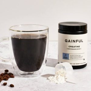 Glass mug of black coffee beside a Gainful Creatine container with spilled creatine powder.