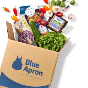 A Blue Apron meal kit box with various fresh ingredients spilling out, including lettuce, red cabbage, garlic, peppers, zucchini, and a package of grass-fed ground beef. A recipe card is also visible, showcasing a prepared meal.