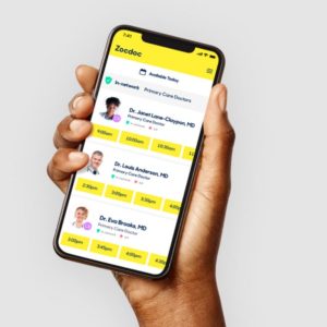 Hand holding a smartphone displaying the Zocdoc app with a list of primary care doctors and available appointment times.