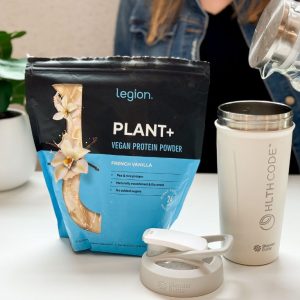 person pouring water into a shaker bottle next to a black and white vanilla flavored legion plant+ vegan protein powder bag