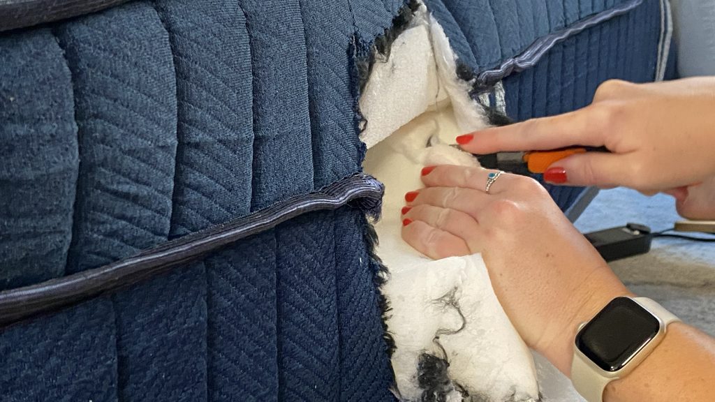 Close-up of a tester using a knife to cut open the side of a blue mattress, revealing the white inner stuffing. The person is wearing a smartwatch and has red nail polish