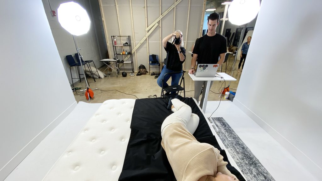 Photo shoot setup for mattress testing, featuring a tester lying on a mattress in a studio with bright lighting and a photographer taking an image of her from the end of the bed. Another tester stands at a laptop, monitoring data or images