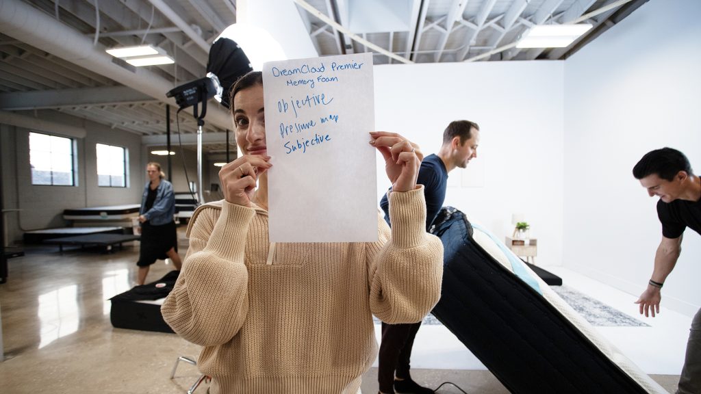 Tester holding up a handwritten sign in a mattress testing studio, with text reading 'DreamCloud Premier Memory Foam, objective, pressure map, subjective.' In the background, two testers are setting up mattresses