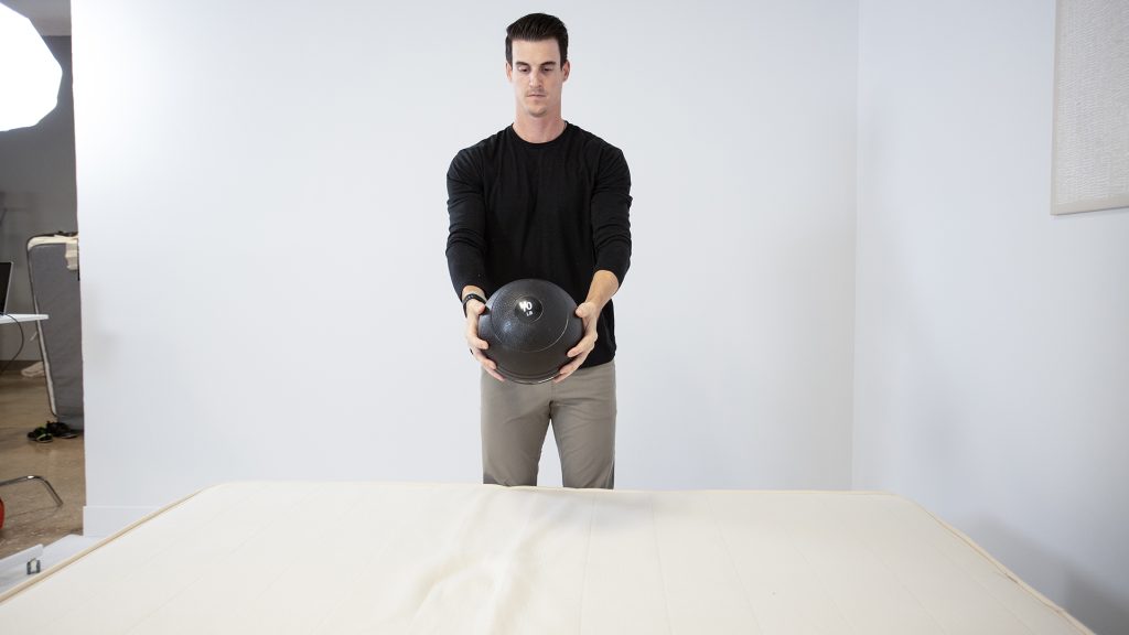 Tester holding a black medicine ball above a mattress, preparing to drop it as part of a mattress firmness test