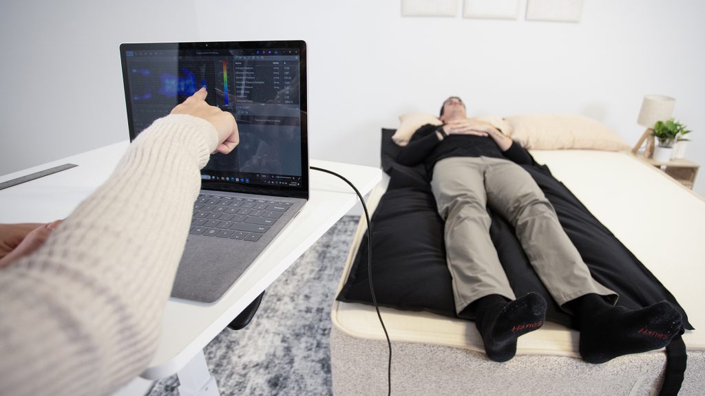 Tester pointing at pressure mapping data displayed on a laptop while another person lies on the map on top of a mattress in the background