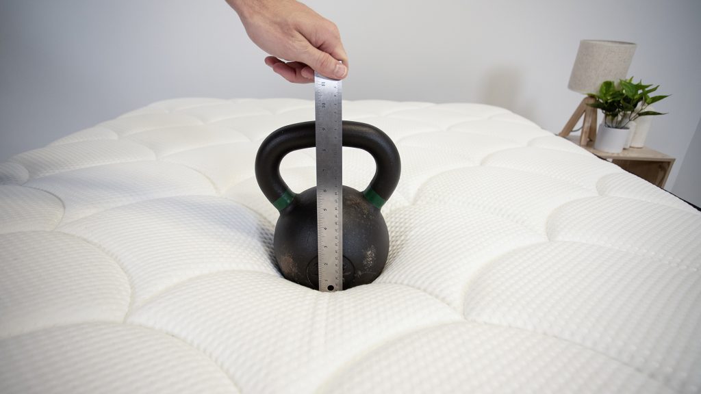 Close-up of a black kettlebell placed on a mattress, with a hand holding a ruler to measure the depth of the mattress indentation