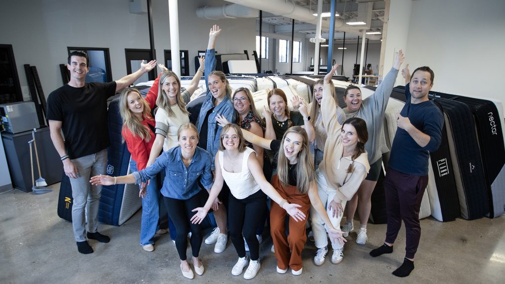 Group of people posing in a bright office space with mattresses stacked in the background after mattress testing. They are standing together with arms raised and open in a cheerful manner