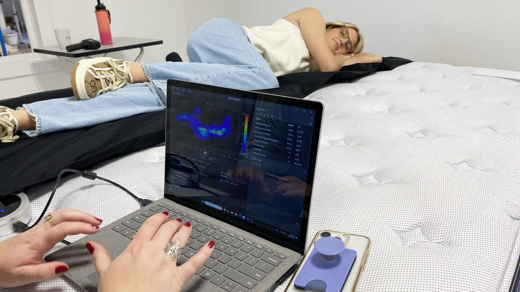 Tester lying on a mattress during a pressure mapping test, with another person typing on a laptop displaying pressure mapping data